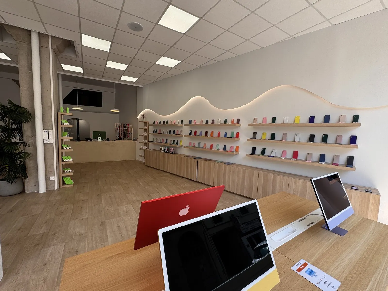 Intérieur de la boutique : mur blanc aux étagères de chêne clair en forme de vague éclairée, coques colorées, iMac sur une table en bois