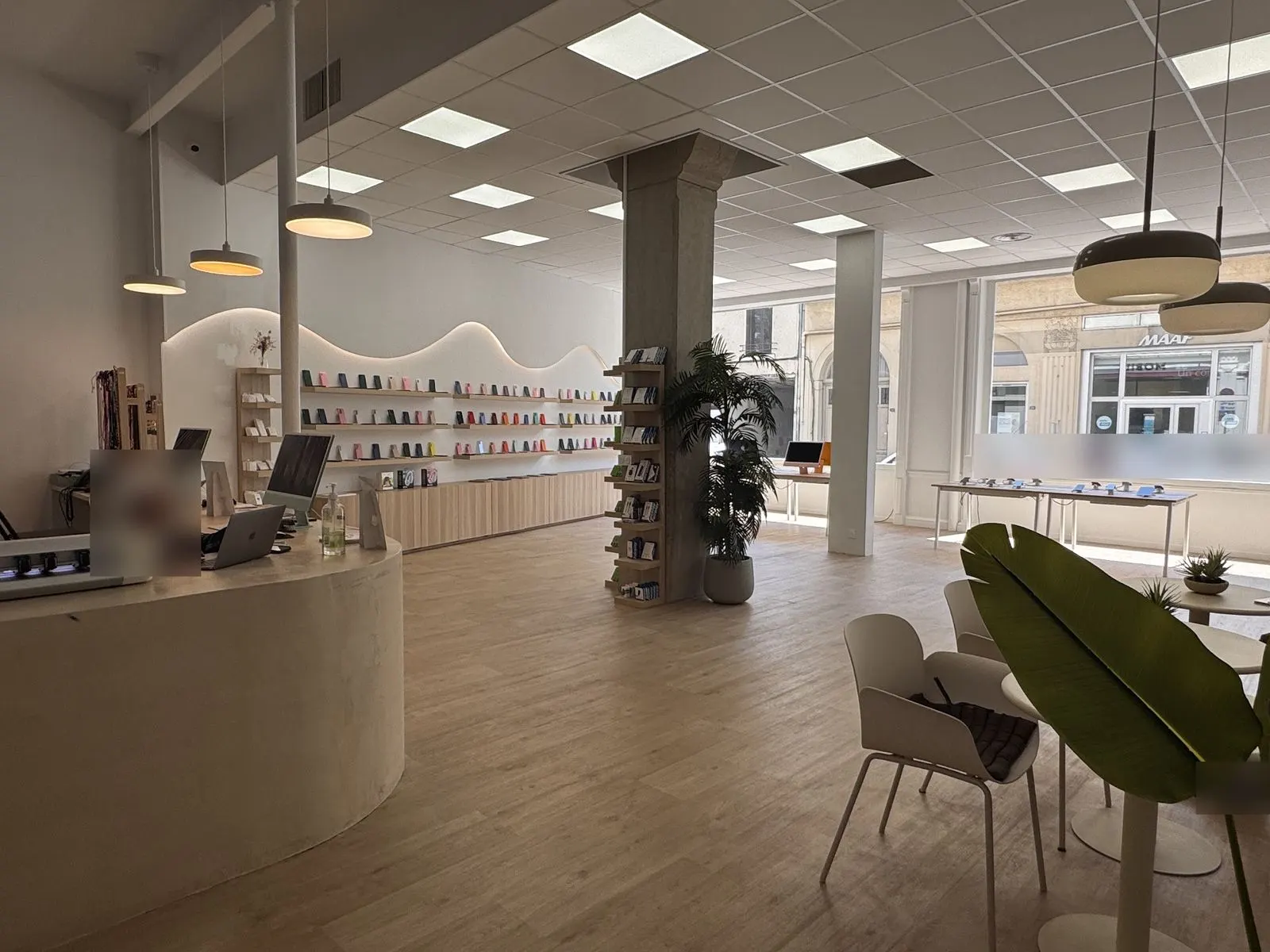 Intérieur de la boutique de Villefranche : comptoir arrondi, sol en bois clair, mur d'étagères en vague éclairée, grande vitrine sur la rue. Personne floutée au comptoir.
