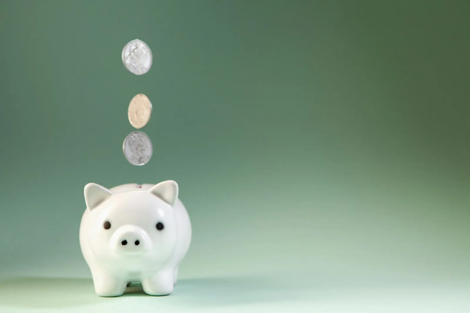 Pièces de monnaie tombant une à une dans une tirelire blanche, fond vert pâle